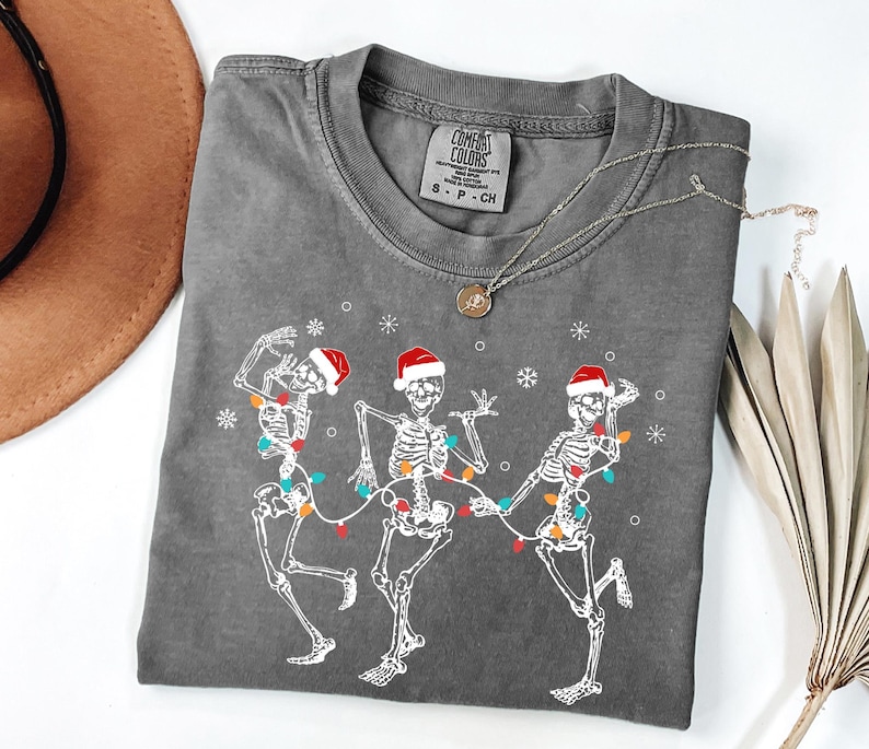 May include: Gray t-shirt featuring a graphic of three dancing skeletons wearing Santa hats and strung with Christmas lights. The skeletons are white, and the lights are colorful. The shirt has a crew neck and short sleeves.