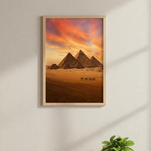 May include: A framed print depicting the pyramids of Giza at sunset. The image shows the ancient structures in a desert setting, with camels in the foreground. The artwork is displayed on a light-coloured wall, creating a warm, inviting atmosphere.