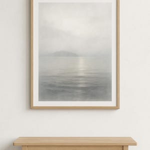 May include: A framed print of a coastal landscape in muted grey tones. The artwork shows a misty seascape with a distant mountain. The print is in a light-coloured wooden frame, and is above a wooden bench.
