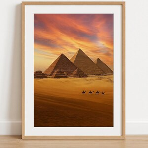 Può includere: Stampa incorniciata con le piramidi di Giza sotto un cielo vibrante al tramonto. L'immagine mostra le piramidi nel deserto con i cammelli. L'opera è racchiusa in una cornice di legno marrone chiaro.