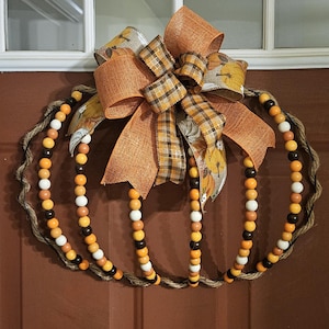 May include: A pumpkin-shaped wreath with a jute rope base and vertical strands of wooden beads in shades of orange, brown, and white. A large bow made of orange burlap and plaid ribbon sits at the top, perfect for fall decor.