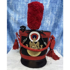 Puede incluir: Un sombrero militar negro y rojo con un emblema de águila dorada y una pluma roja. El sombrero tiene un emblema circular blanco y azul con una corona en la parte superior. El sombrero está decorado con un cordón rojo y borlas.