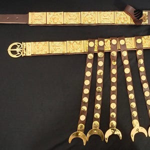 May include: A brown leather belt with a gold metal buckle and decorative plates. The belt has five leather straps with gold metal studs and crescent-shaped ends.