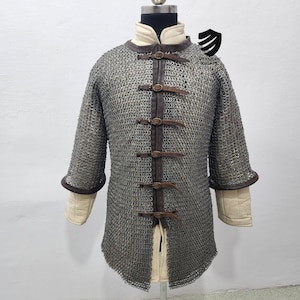 Könnte beinhalten: Ein silbernes Kettenhemd mit braunen Lederriemen und Schnallen. Das Hemd hat lange Ärmel und einen spitzen Saum. Das Kettenhemd besteht aus kleinen, ineinandergreifenden Metallringen.