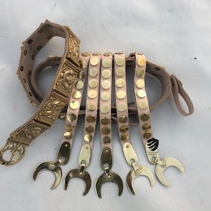 May include: A gold-colored leather belt with decorative metal accents and five straps with round metal studs. The belt is made of leather and has a buckle. The straps are attached to the belt with metal loops. The belt is a replica of a Roman military belt.
