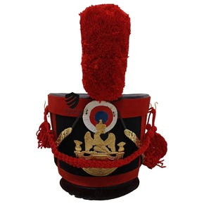 Puede incluir: Un sombrero militar negro y rojo con un emblema de águila dorada y una pluma roja. El sombrero tiene una banda negra con una trenza dorada y un borlón rojo.