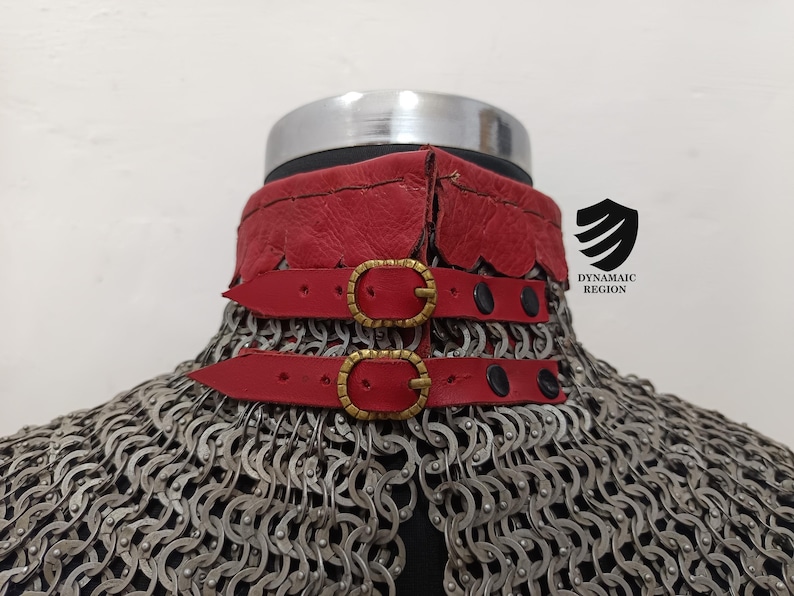 May include: A red leather collar with two gold buckles and black straps. The collar is attached to a chainmail garment.