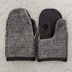 Puede incluir: Un par de guantes de malla con palmas y puños de cuero. Los guantes están hechos de anillos de metal entrelazados y tienen un puño de cuero negro con el texto "Dynamac Region" en él.