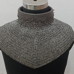 Può includere: Un collare di maglia di colore grigio con un colletto in pelle marrone. La maglia è realizzata con piccoli anelli di metallo intrecciati.