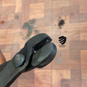 May include: Close-up of a black metal tool with a small hole in the top. The tool is held in a hand and the background is a wooden surface.