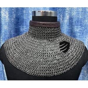 May include: A silver chainmail collar with a black leather trim and a black emblem. The chainmail is made of small, interlocking metal rings. The collar is displayed on a black mannequin.