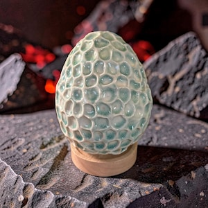 May include: A ceramic sculpture with a textured, honeycomb-like pattern in shades of teal and white. The egg-shaped art piece sits on a small, round wooden base, showcasing a unique design. The sculpture is a decorative item.