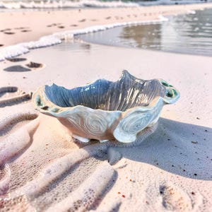 Może przedstawiać: Ceramiczna miska w kształcie muszli, w odcieniach niebieskiego, białego i zielonego. Misa stoi na piaszczystej plaży, z morzem w tle. Średnica miski wynosi około 15 cm.