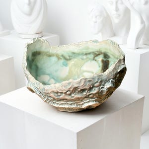 Pode incluir: Uma tigela de cerâmica artesanal com um exterior texturizado e um interior liso e esmaltado. A tigela apresenta uma forma orgânica única com uma paleta de cores azul claro, verde e castanho. A tigela é exibida numa superfície branca.