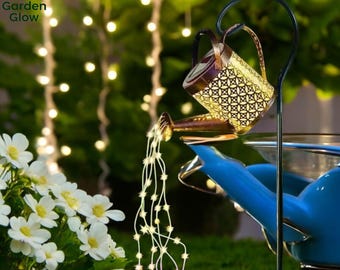 May include: A decorative metal watering can with a light string of lights pouring out of the spout. The watering can is gold with a patterned design and is sitting on a blue watering can. The background is blurred and shows a string of lights.