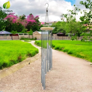 Può includere: Un carillon d'argento con più tubi appesi a un anello di legno. Il carillon è sospeso da un gancio di metallo nero e pende in un giardino.
