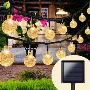 Op de afbeelding: Een string met solar-aangedreven gloeilampen met een zonnepaneel. De lampen zijn wit en hebben een matglazen afwerking. De lampen zijn aan een zwarte draad bevestigd en hangen aan een tak.