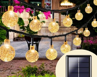 Puede incluir: Una cadena de luces solares en forma de globo con un panel solar. Las luces son blancas y tienen un acabado de vidrio esmerilado. Las luces están enhebradas en un cable negro y están colgadas de una rama de árbol.