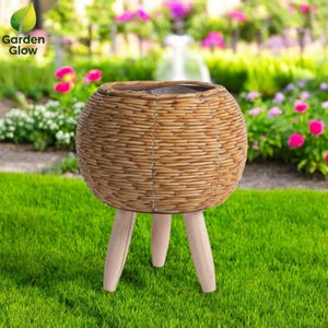 Puede incluir: Un macetero redondo de mimbre marrón con tres patas de madera. El macetero está sobre una superficie de hierba verde.