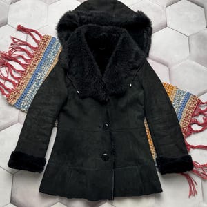 May include: A black coat with a hood and faux fur trim. The coat features a button-up front, long sleeves, and a flared hem. The hood and collar are lined with black faux fur. The coat is displayed on a patterned surface.
