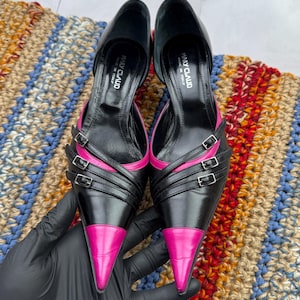 May include: A pair of black leather high heels with pink leather accents. The heels have multiple straps with buckles and pointed toes. The shoes are made in Italy by the brand "Many Claud".