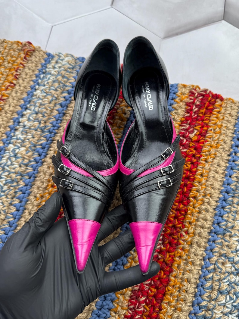 May include: A pair of black leather high heels with pink leather accents. The heels have multiple straps with buckles and pointed toes. The shoes are made in Italy by the brand "Many Claud".