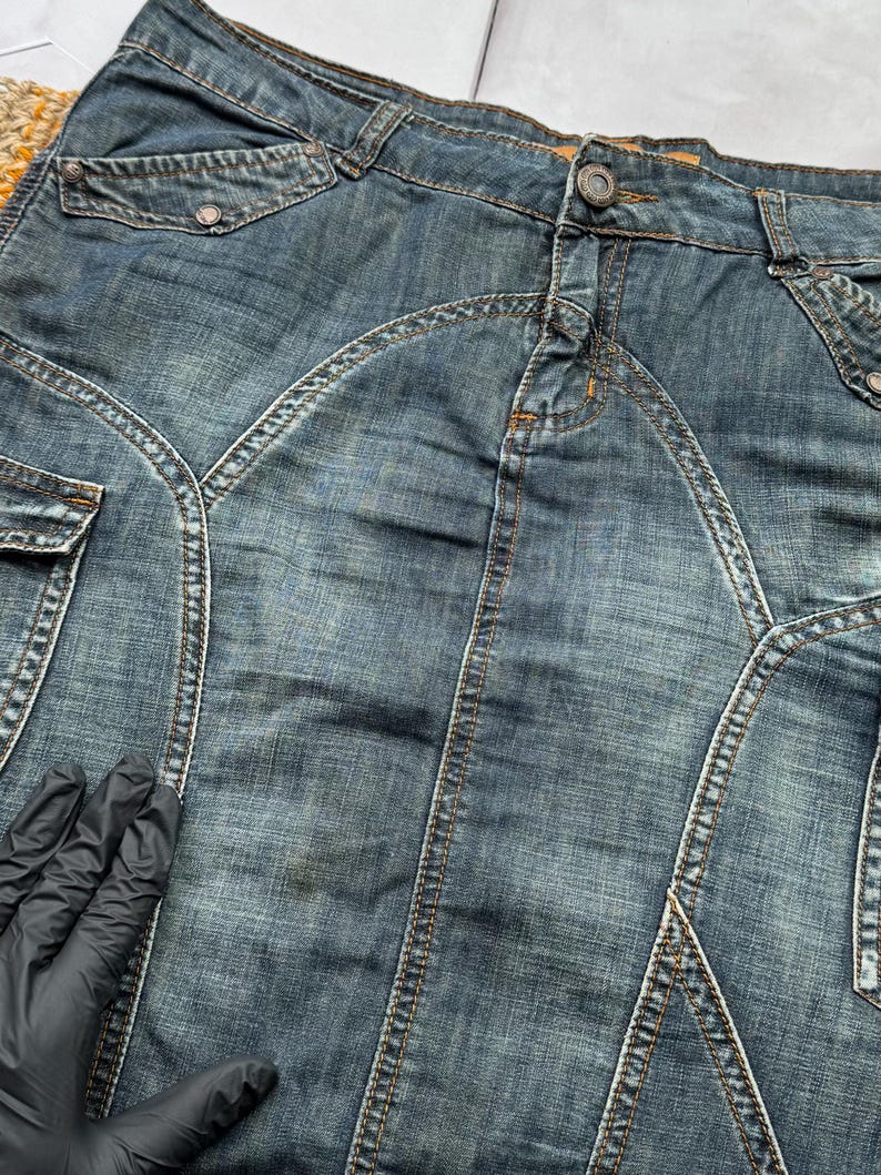 Puede incluir: Una falda vaquera en un lavado medio con costuras visibles y detalles de bolsillo. La falda tiene cierre de bot&oacute;n y un dise&ntilde;o de costura &uacute;nico. La tela muestra signos de desgaste, lo que aumenta su atractivo vintage. Una prenda de vestir cl&aacute;sica.