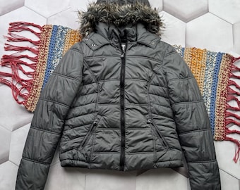 May include: A gray puffer jacket with a faux fur-trimmed hood. The jacket has a quilted design, a zipper closure, and two front pockets. The jacket is displayed on a colorful woven textile.