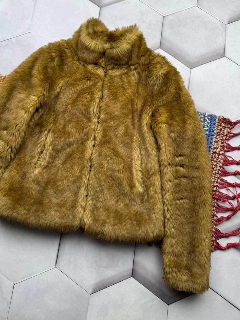 May include: A soft, plush faux fur jacket in a muted olive green color. The jacket has a high collar and a zipper closure.