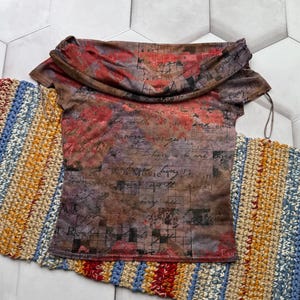 Op de afbeelding: Een top met korte mouwen en een gedrapeerde halslijn. De top heeft een print met rode, bruine en paarse bloemen- en scriptontwerpen. Het kledingstuk wordt gepresenteerd op een kleurrijke, gestreepte, gehaakte loper.