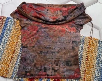 May include: A short-sleeved top with a draped neckline. The top features a print with red, brown, and purple floral and script designs. The garment is displayed on a colorful, striped, crocheted runner.