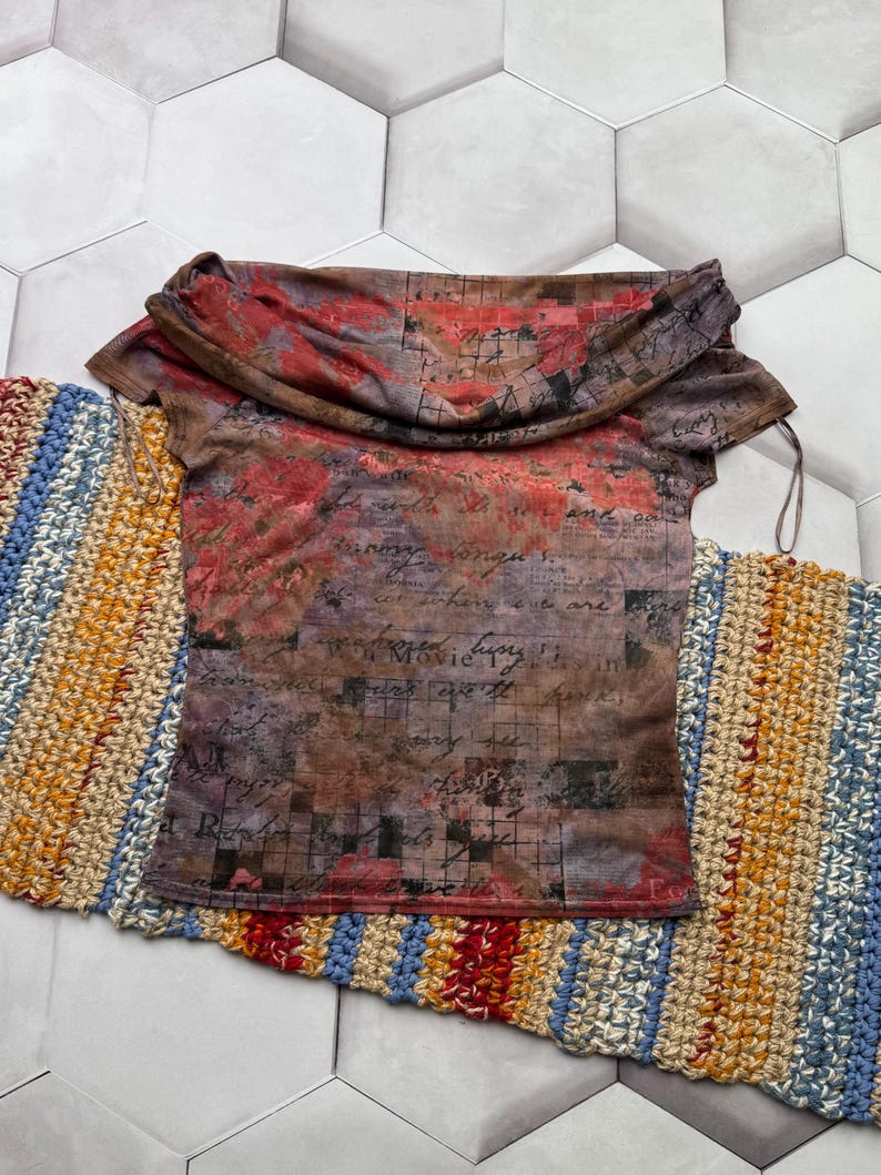 Op de afbeelding: Een top met korte mouwen en een gedrapeerde halslijn. De top heeft een print met rode, bruine en paarse bloemen- en scriptontwerpen. Het kledingstuk wordt gepresenteerd op een kleurrijke, gestreepte, gehaakte loper.