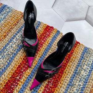 May include: A pair of black and pink high heel shoes with a pointed toe and buckle accents. The shoes are on a striped rug.
