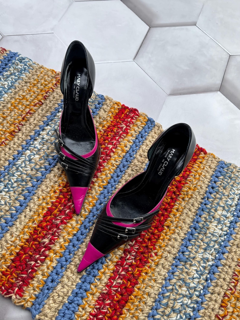 May include: A pair of black and pink high heel shoes with a pointed toe and buckle accents. The shoes are on a striped rug.