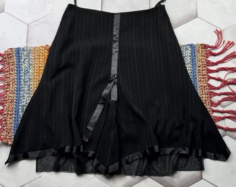 Puede incluir: Falda plisada negra con un detalle de cinta de satén en la parte delantera y un dobladillo de satén. La falda tiene una cintura alta y una silueta acampanada. La falda cuelga sobre un fondo de baldosas hexagonales blancas y grises.