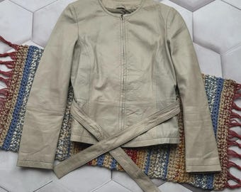 Könnte beinhalten: Hellbeige Lederjacke mit rundem Kragen und Reißverschluss vorne. Die Jacke hat lange Ärmel und einen passenden Gürtel, der um die Taille gebunden wird. Die Jacke wird auf einem farbenfrohen gewebten Teppich präsentiert.