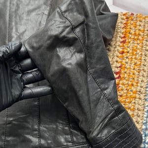 K&ouml;nnte beinhalten: Nahaufnahme einer schwarzen Lederjacke. Die Jacke hat detaillierte N&auml;hte an den N&auml;hten, Manschetten und am Bund. Das Leder wirkt abgenutzt, mit sichtbarer Textur und subtilen Farbvariationen. Der &Auml;rmel ist gebogen.