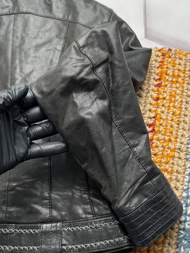 K&ouml;nnte beinhalten: Nahaufnahme einer schwarzen Lederjacke. Die Jacke hat detaillierte N&auml;hte an den N&auml;hten, Manschetten und am Bund. Das Leder wirkt abgenutzt, mit sichtbarer Textur und subtilen Farbvariationen. Der &Auml;rmel ist gebogen.