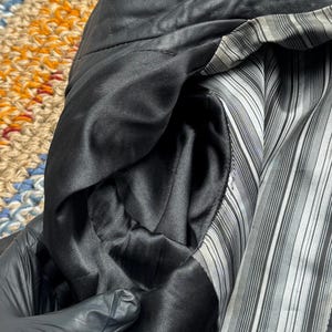 K&ouml;nnte beinhalten: Nahaufnahme des Innenraums einer schwarzen Lederjacke. Das Futter ist ein schwarz-wei&szlig; gestreifter Stoff. Die Tasche ist mit einem glatten, schwarzen Satinmaterial gef&uuml;ttert. Der Kragen der Jacke ist sichtbar.