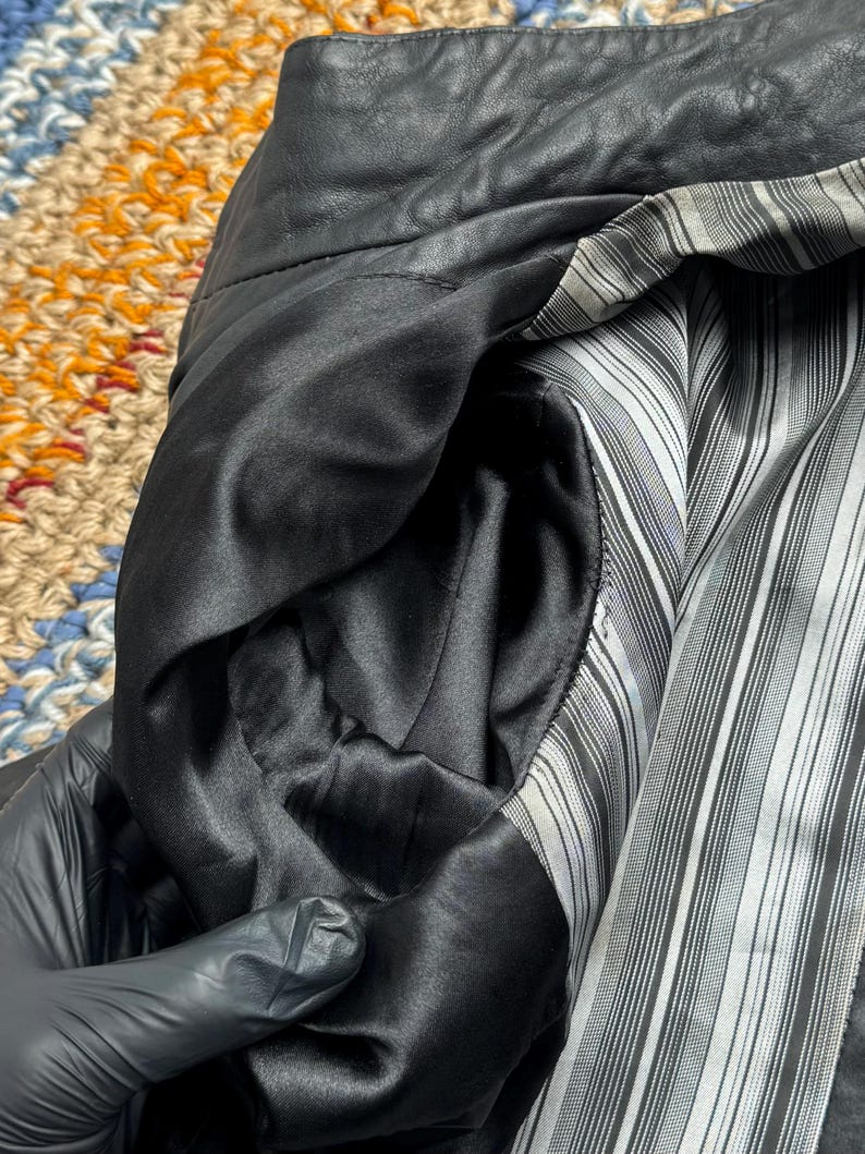 K&ouml;nnte beinhalten: Nahaufnahme des Innenraums einer schwarzen Lederjacke. Das Futter ist ein schwarz-wei&szlig; gestreifter Stoff. Die Tasche ist mit einem glatten, schwarzen Satinmaterial gef&uuml;ttert. Der Kragen der Jacke ist sichtbar.