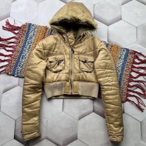 May include: A cropped, gold-colored puffer jacket with a hood lined with faux fur. The jacket features two front pockets, a ribbed waistband, and a zipper closure. A colorful, fringed scarf is partially visible.