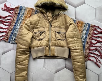 May include: A cropped, gold-colored puffer jacket with a hood lined with faux fur. The jacket features two front pockets, a ribbed waistband, and a zipper closure. A colorful, fringed scarf is partially visible.