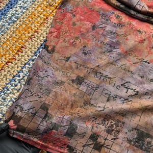 Op de afbeelding: Een doorschijnende top met korte mouwen met een bloemen- en scriptprint in tinten rood, paars en bruin. De stof heeft een mesh-achtige textuur. De top is gedrapeerd over een veelkleurig, gehaakt oppervlak.
