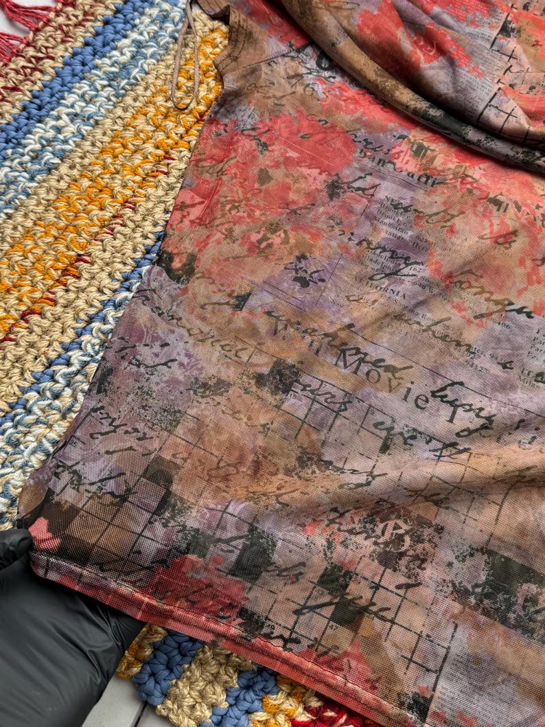 Op de afbeelding: Een doorschijnende top met korte mouwen met een bloemen- en scriptprint in tinten rood, paars en bruin. De stof heeft een mesh-achtige textuur. De top is gedrapeerd over een veelkleurig, gehaakt oppervlak.