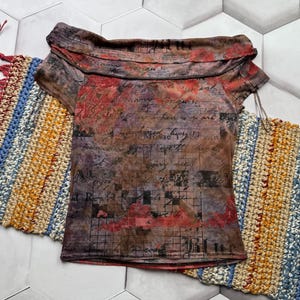 Op de afbeelding: Een top met korte mouwen met een uniek off-the-shoulder ontwerp. De stof heeft een bruin, rood en paars bloemen- en scriptpatroon. De top wordt gepresenteerd op een wit en grijs zeshoekig tegeloppervlak, met kleurrijke haakartikelen op de achtergrond.