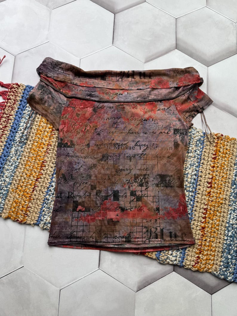 Op de afbeelding: Een top met korte mouwen met een uniek off-the-shoulder ontwerp. De stof heeft een bruin, rood en paars bloemen- en scriptpatroon. De top wordt gepresenteerd op een wit en grijs zeshoekig tegeloppervlak, met kleurrijke haakartikelen op de achtergrond.