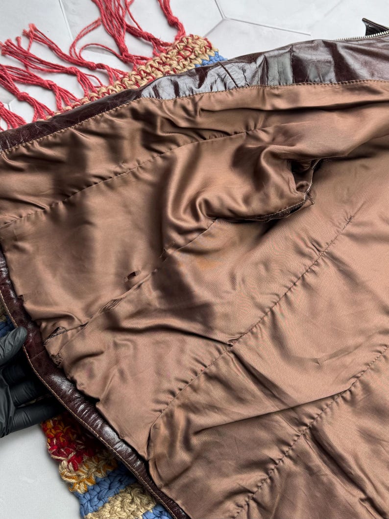 Pu&ograve; includere: L'interno di una giacca di pelle marrone con una fodera in raso marrone. La giacca &egrave; appoggiata su una coperta all'uncinetto colorata.