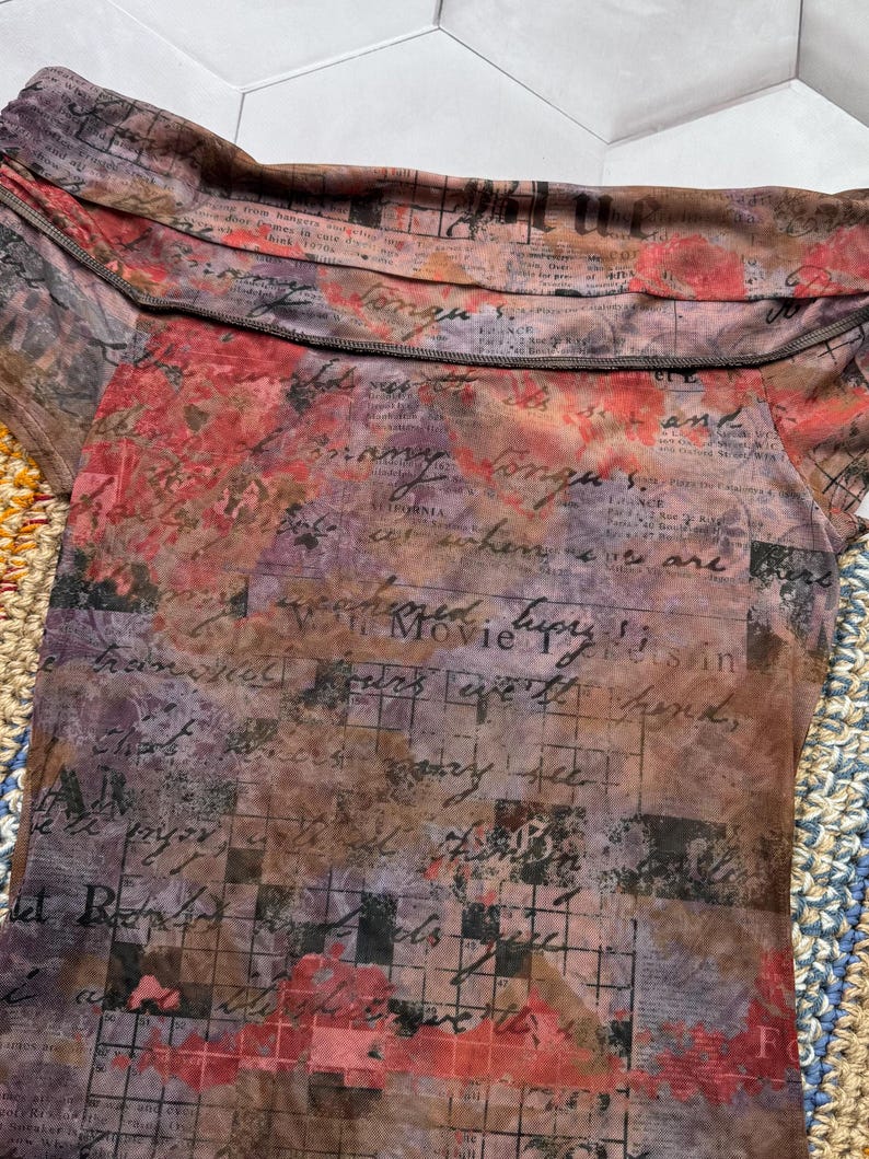 Op de afbeelding: Een doorschijnende off-the-shoulder top met een unieke print. De top heeft een mix van kleuren, waaronder bruin, rood en paars, met een krantenachtig ontwerp en handgeschreven tekst. De stof heeft een zachte, vloeiende uitstraling.
