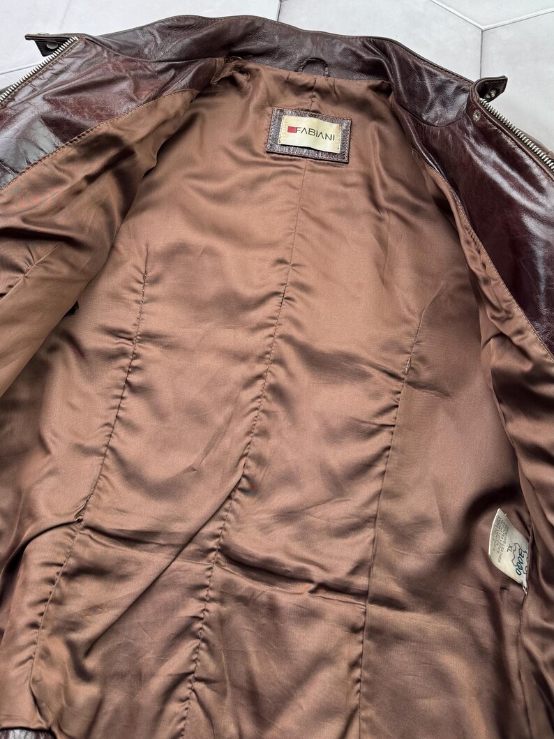 Pu&ograve; includere: Giacca di pelle marrone con chiusura a zip e un'etichetta che recita "FABIANI". La giacca &egrave; non foderata e ha una fodera in raso marrone.