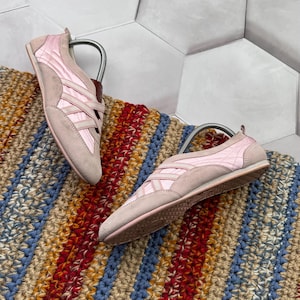 Peut inclure: Une paire de baskets rose clair avec un bout en daim et un design matelassé sur les côtés. Les chaussures sont présentées sur un tapis rayé coloré. Les baskets ont une semelle en caoutchouc et un détail en cuir marron au talon.